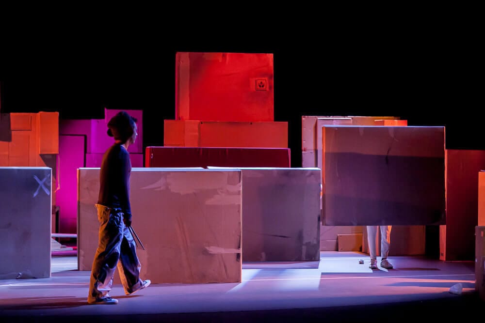 Intérprete de circo detrás de una caja junto a la escenografía de cajas en una escena de Ludo.